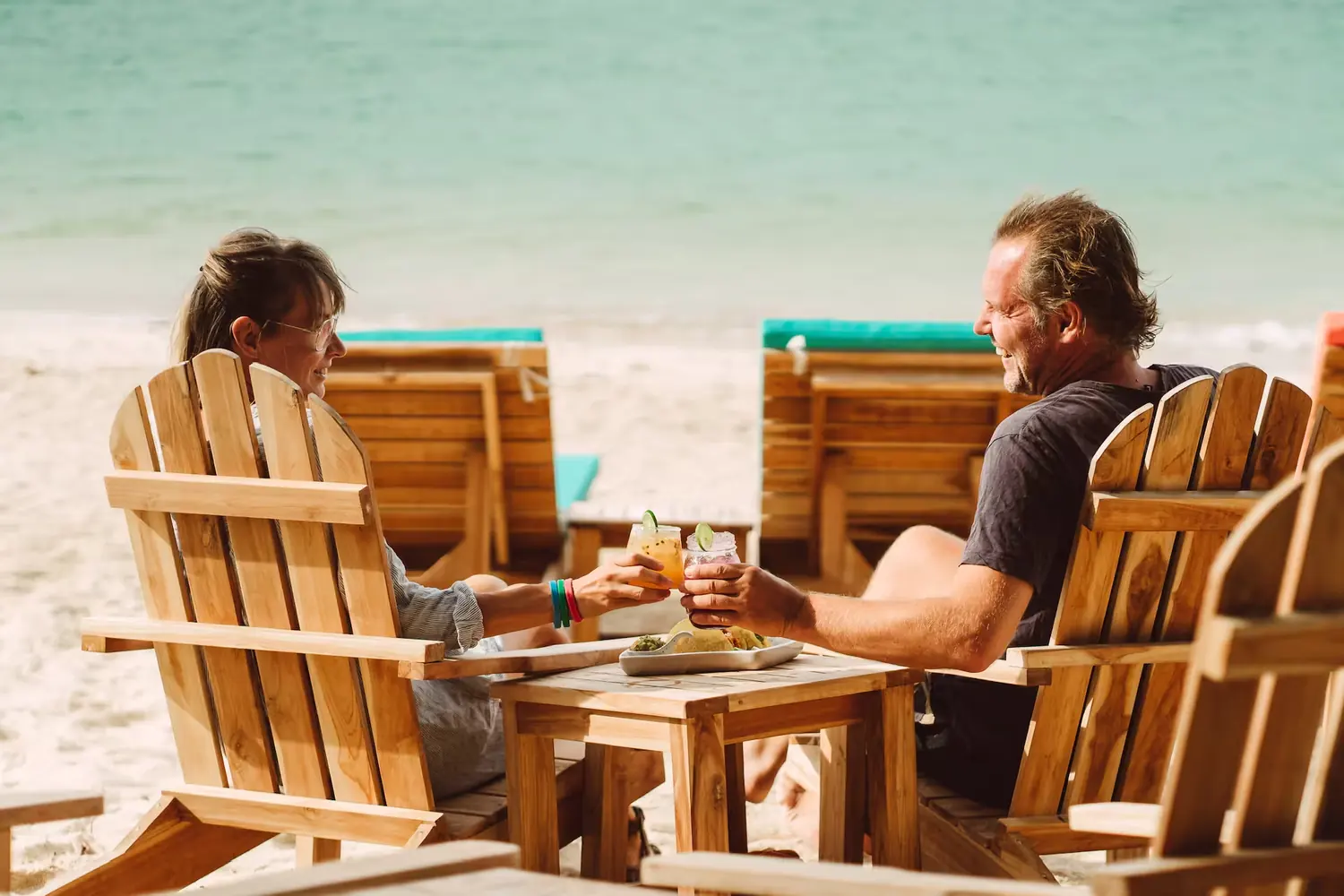 Two people in wooden beach chairs on the sand, raising drinks over a shared plate of food.