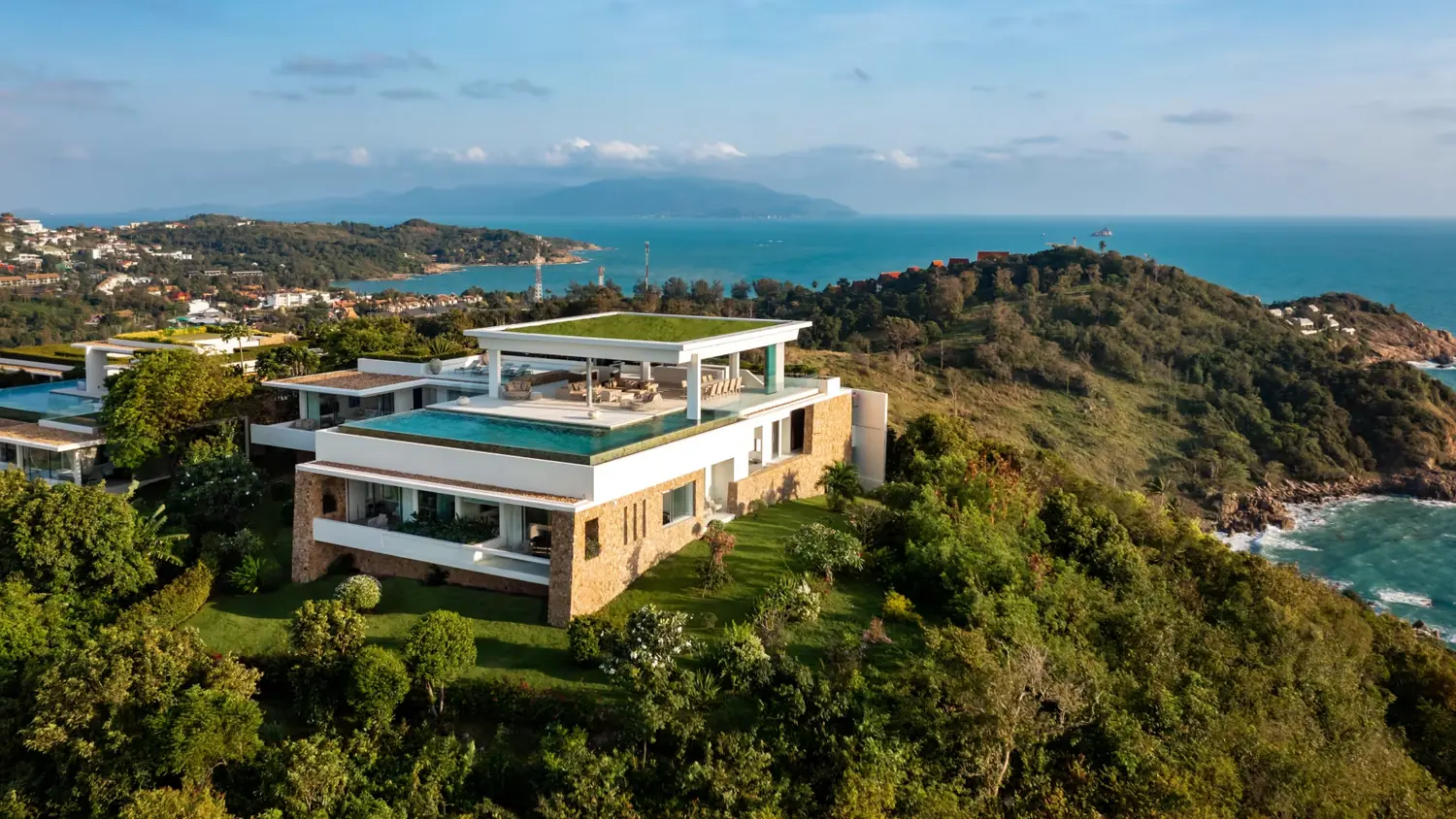An aerial view of a modern hillside villa with an infinity pool, above a green headland and the sea.